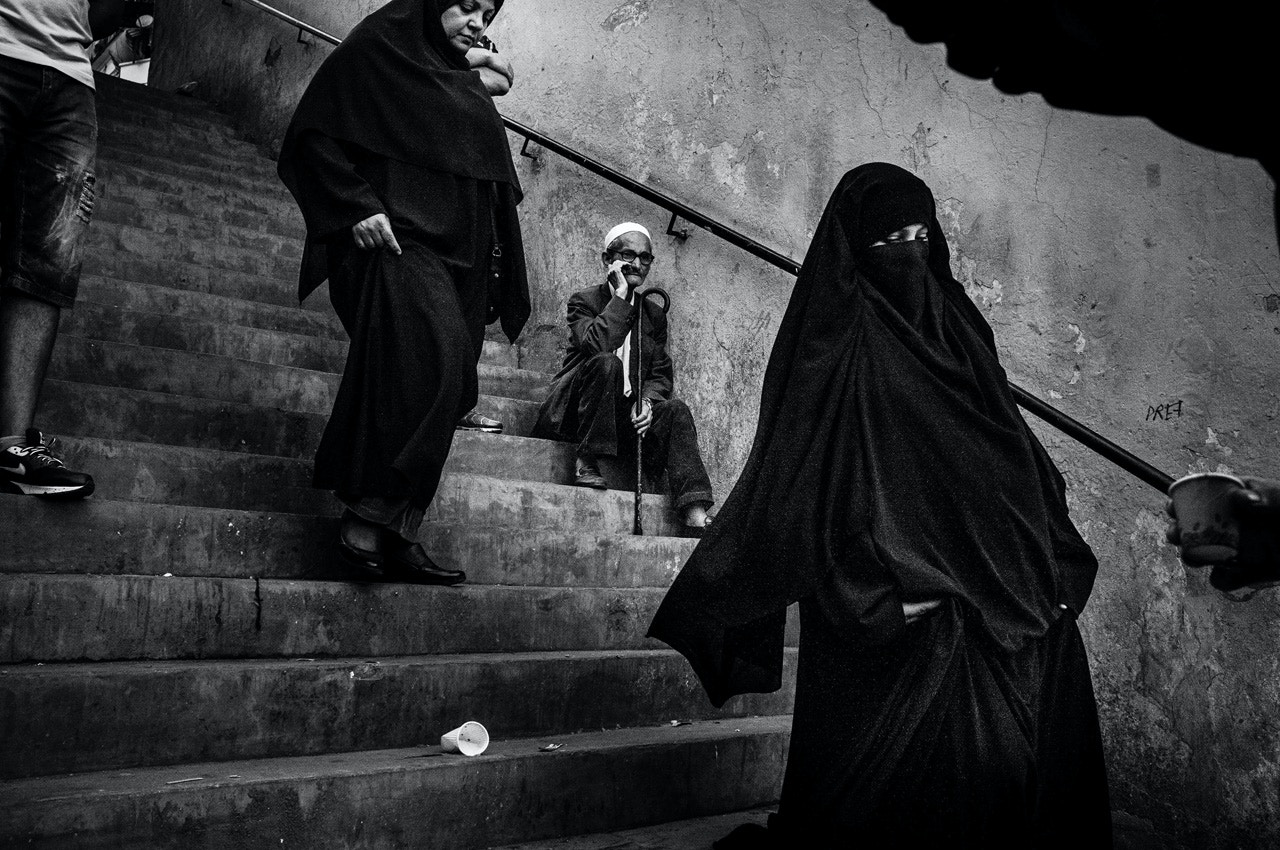 2014年5月31日，阿爾及利亞阿爾及爾（Algiers）巴布瓦德 (Bab el-Oued)，一名老人坐在梯級上，旁邊戴着尼卡布面紗（Niqab）的女士經過。近20年，尼卡布面紗在巴布瓦德愈來愈普及。年度圖片故事及長期計劃（故事）冠軍。（Romain Laurendeau）
