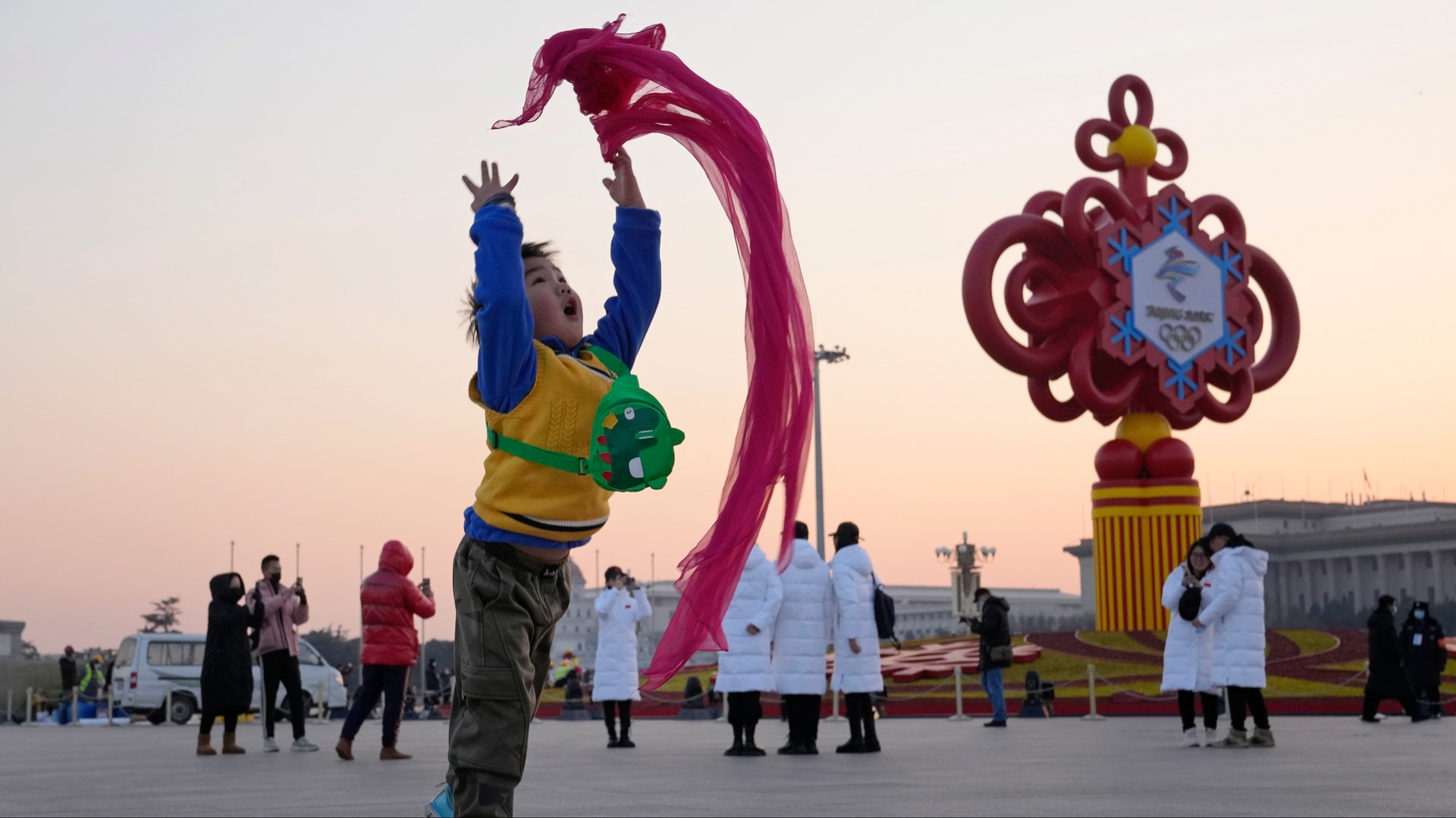 圖為2022年1月18日，一名兒童在北京天安門廣場拋起一條紅色的圍巾。他的後方是北京冬奧的戶外大型裝飾裝置。（AP）
