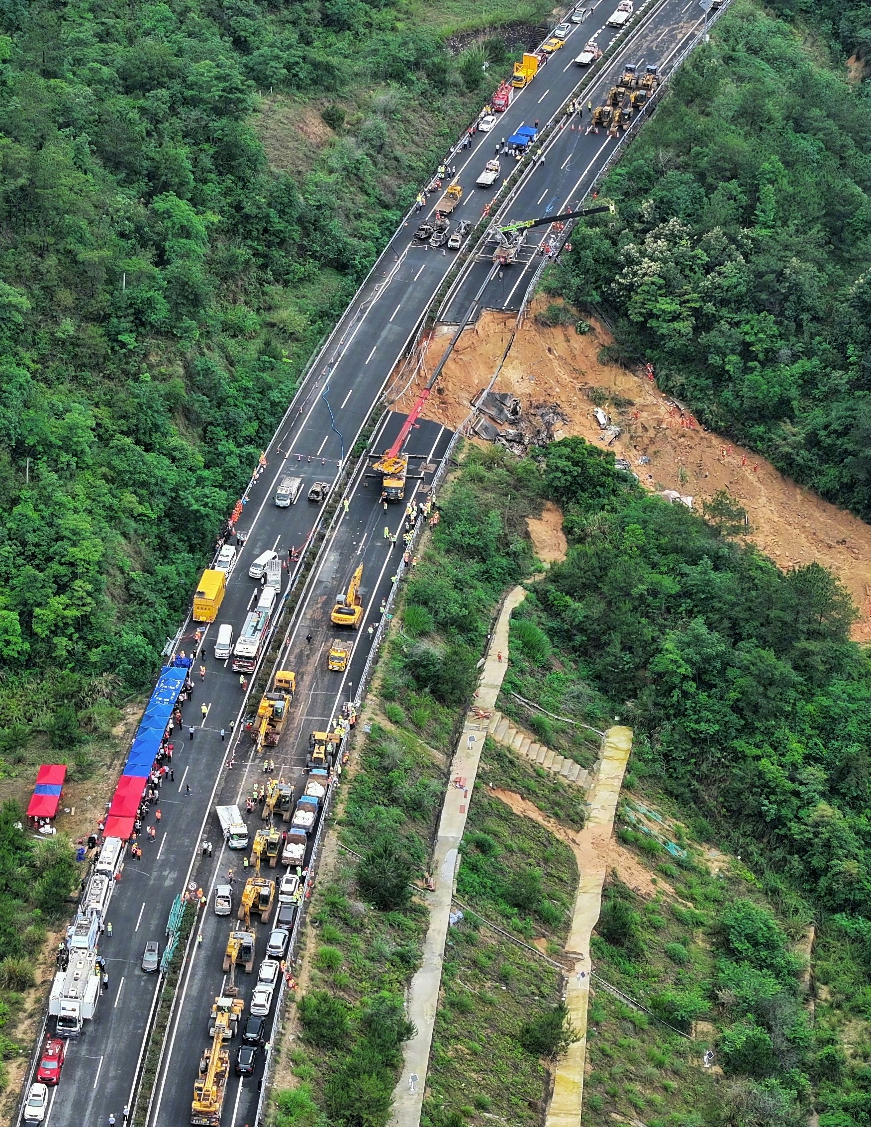 新华社发布事发现场的高清航拍图，可以清楚看见高速公路上的路陷范围，该处路面整个向山坡下垮了下去，泥土滑出至少几十米远，只剩下高速护栏连著桩柱悬空在那里。塌陷的位置形成了一个大坑，内有大量被烧毁的汽车。（新华社）