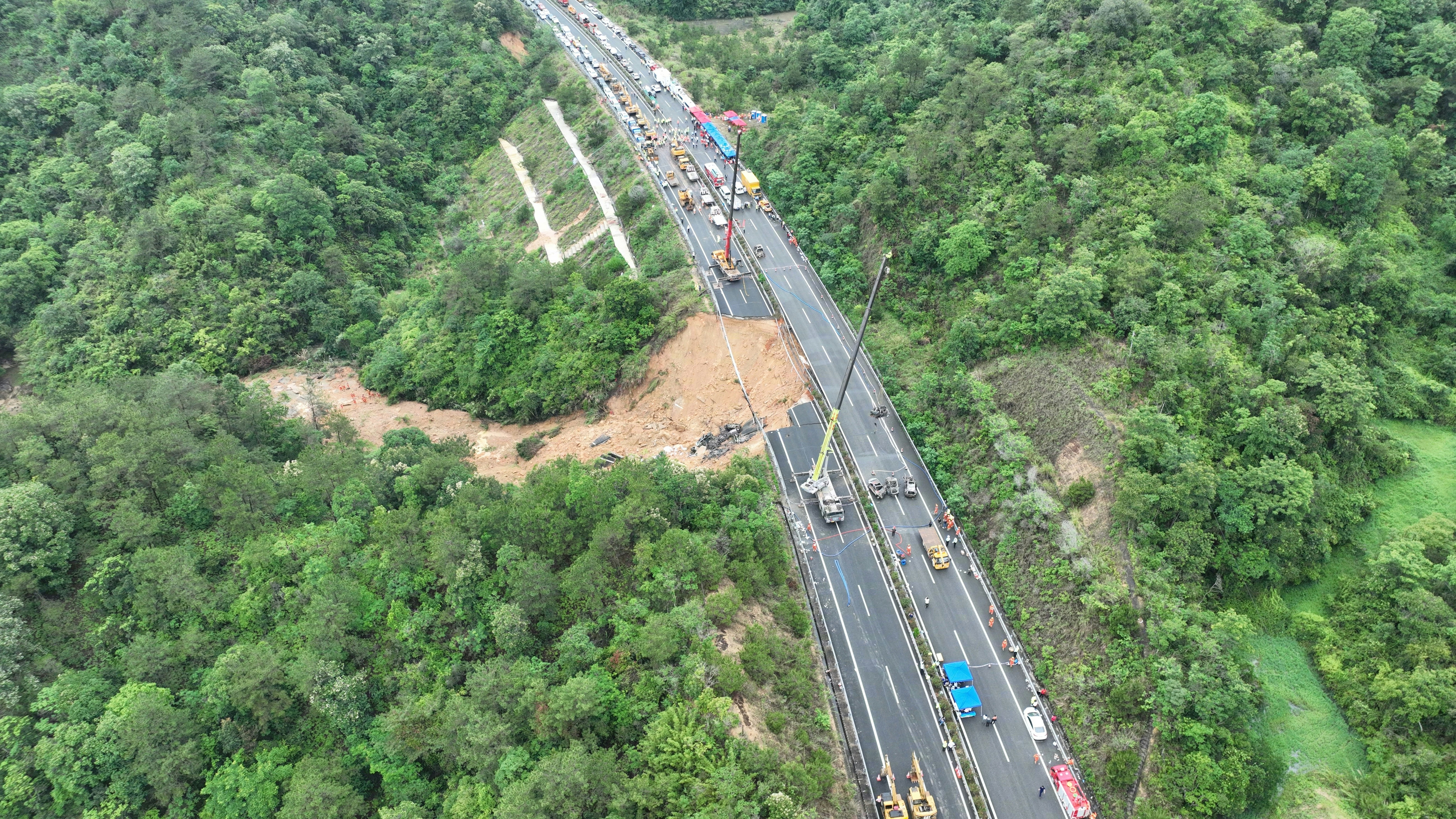 新华社发布事发现场的高清航拍图，可以清楚看见高速公路上的路陷范围，该处路面整个向山坡下垮了下去，泥土滑出至少几十米远，只剩下高速护栏连著桩柱悬空在那里。塌陷的位置形成了一个大坑，内有大量被烧毁的汽车。（新华社）