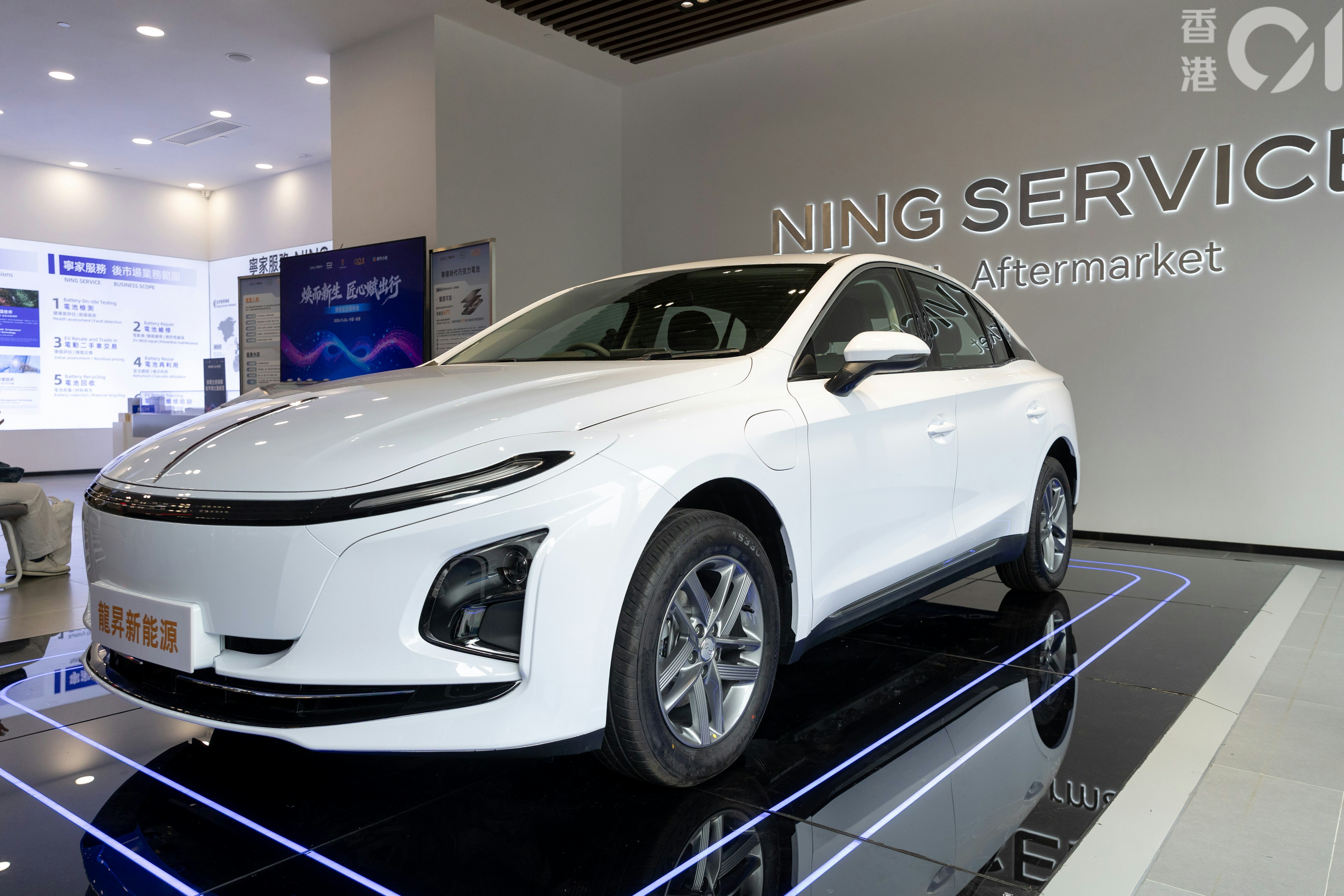 A white electric car parked indoors at a display featuring the red flag E-QM5, highlighting rapid 99-second battery swapping technology by Dragon Sheng New Energy for electric taxis in Hong Kong.