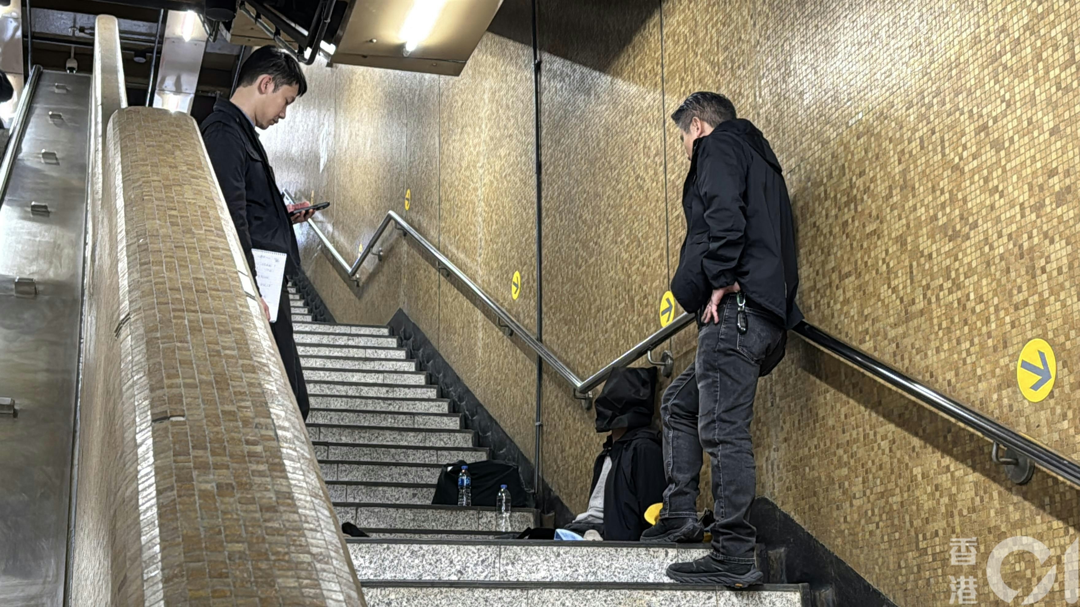被捕人蒙黑色頭套一左一右坐在上環地鐵站B出口樓梯，被多名警員看守。（翁鈺輝攝）