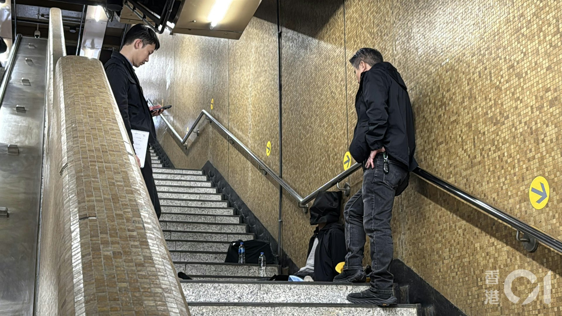 兩名被捕人蒙黑色頭套一左一右坐在上環地鐵站B出口樓梯,被多名警員看守。(翁鈺輝攝)