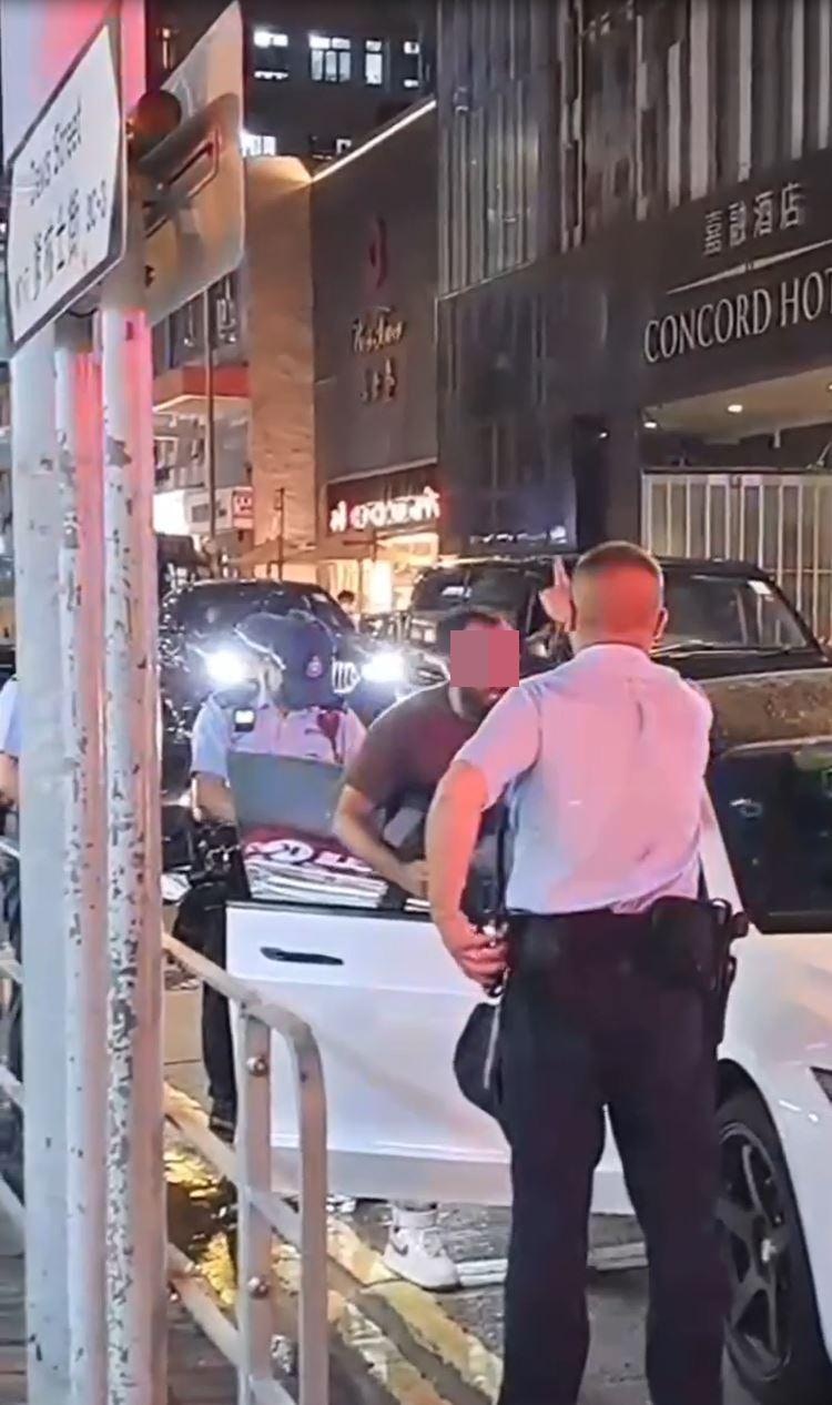非華裔男子在該輛Tesla旁邊一邊舉起手機，一邊操不純正廣東話爆粗表示「我落咗車」、「我揸緊車」、「X你老X」等，警員保持克制，回應指「大家咁話」。（網上影片截圖）