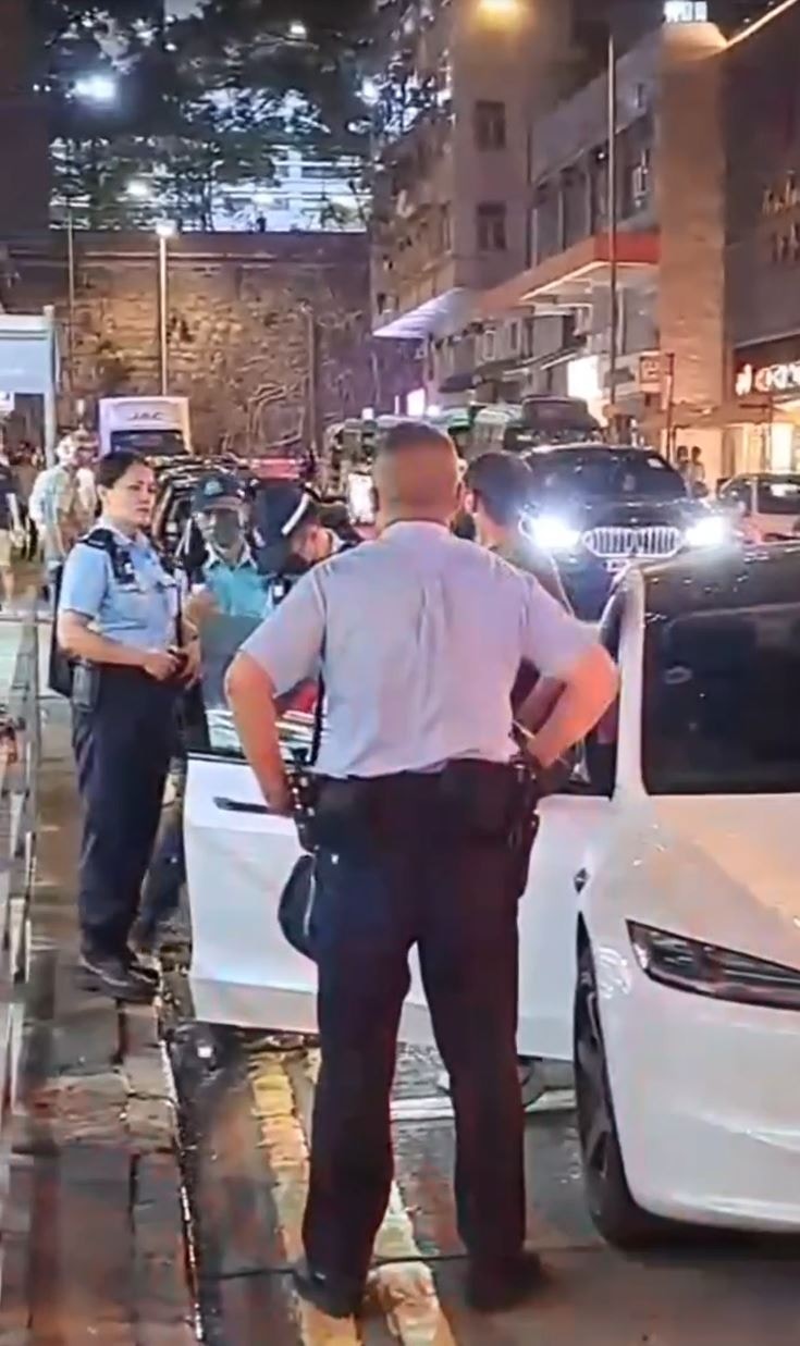 非華裔男子在該輛Tesla旁邊一邊舉起手機，一邊操不純正廣東話爆粗表示「我落咗車」、「我揸緊車」、「X你老X」等，警員保持克制，回應指「大家咁話」。（網上影片截圖）