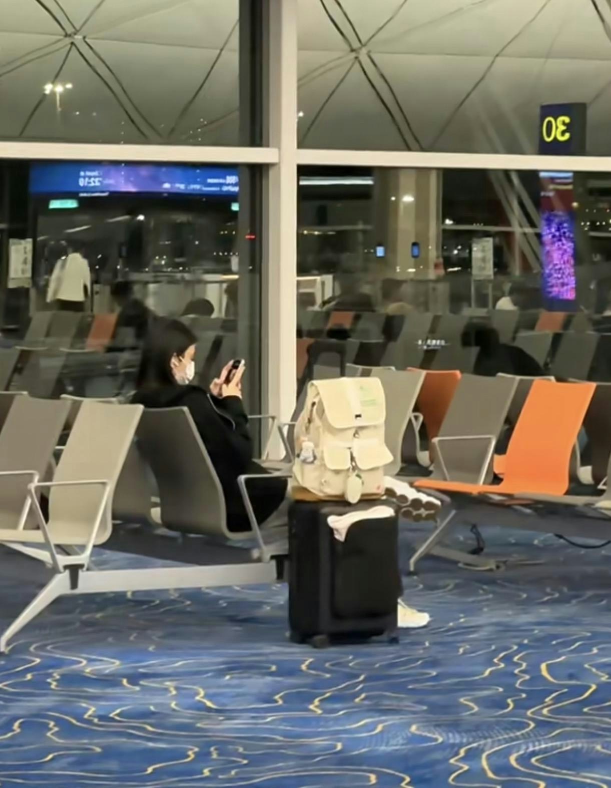 香港の空港で、奚夢瑤がスマートフォンを手に座り、白いリュックとスーツケースをそばに置いている